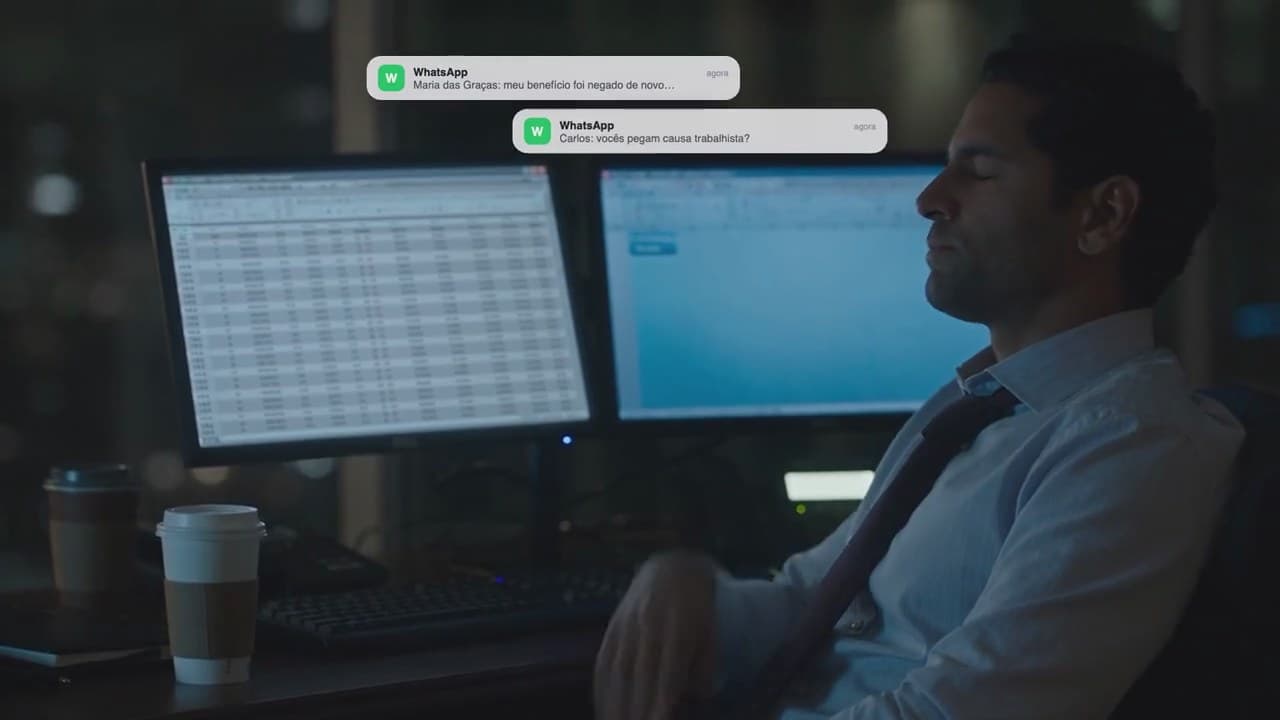 Advogado à noite no escritório, diante de dois monitores, com notificações de WhatsApp se acumulando